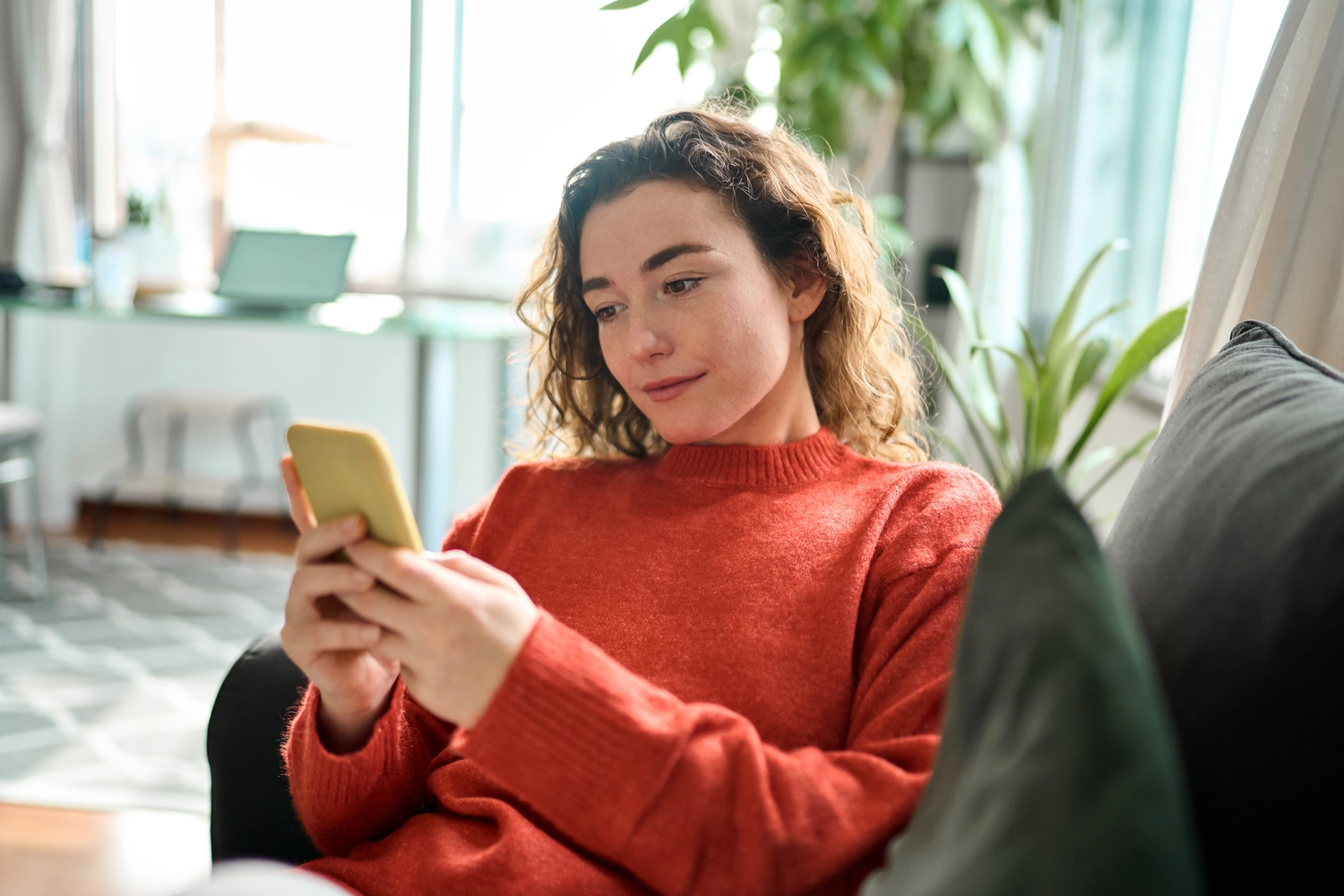 HZ Loop - Tauschapp mit KI - Eine entspannte junge Frau sitzt auf dem Sofa und nutzt ihr Handy. Die glückliche Dame hält ihr Smartphone in der Hand, scrollt, schaut auf ihr Handy und genießt es, online einzukaufen und in mobilen KI-Apps zu chatten.